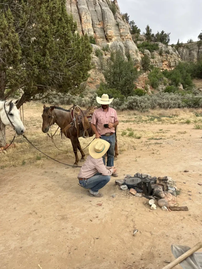 Two cowboys with horses are gathered around a campfire in a desert landscape.